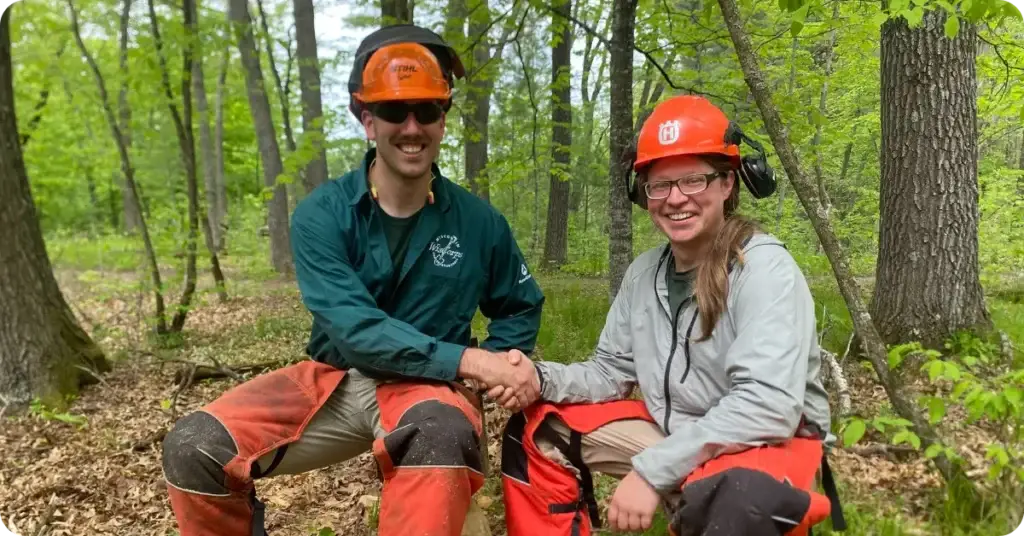 Two WisCorps crew members in hard hats and chainsaw chaps smiling while seated in the woods