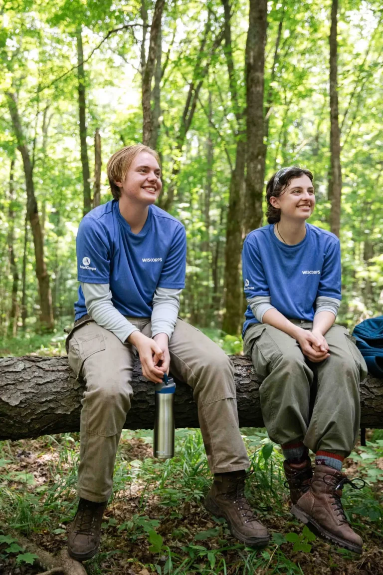 Two WisCorps crew members in blue shirts sitting on a log in a green forest, smiling