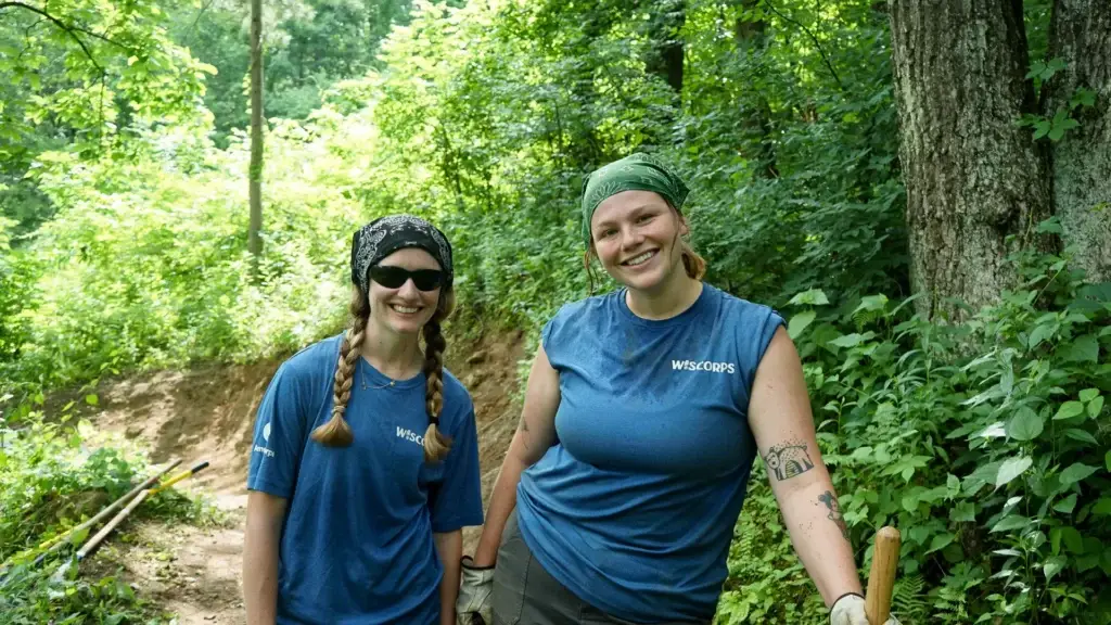 Two smiling WisCorps AmeriCorps crew members in blue WisCorps shirts on a forest trail